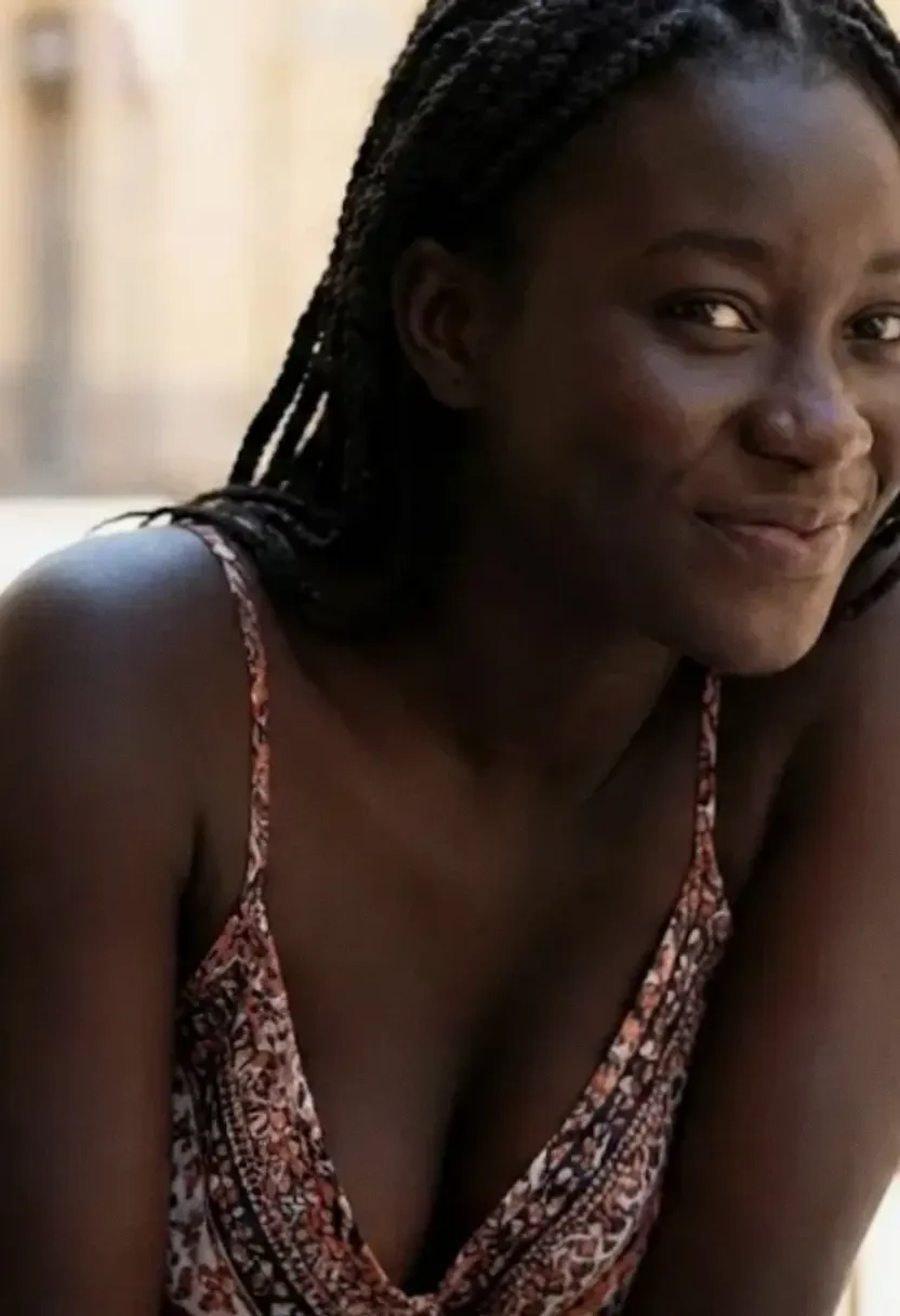 Young Black woman with braided hair seated outdoors with blurred tables and chairs behind her.