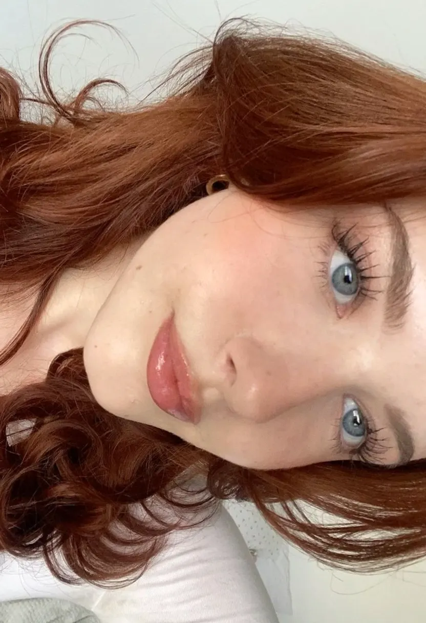 Young woman with long red hair and blue eyes wearing a white top against a light wall.