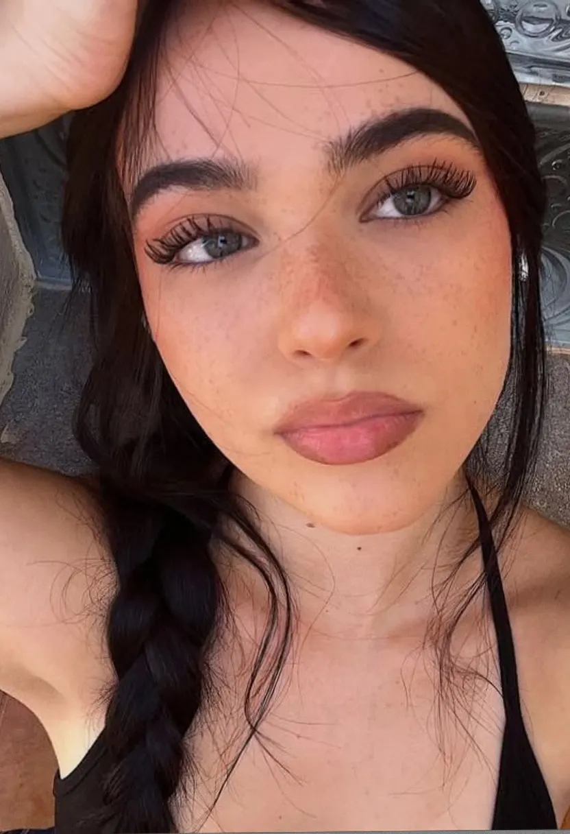 Young woman with long dark braided hair and light green eyes taking a close-up selfie near a textured wall.